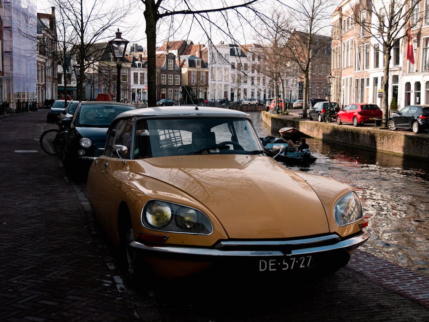 A vintage beige convertible car with a black soft top is parked on a cobbled street beside a canal in a residential area of Holland Park, London. The car's front is facing the camera, with its headlights and license plate clearly visible. Behind the convertible, there is a dark-colored hatchback parked parallel to it, with a bicycle secured upright on its rear rack. The street features traditional terraced houses with large windows and brick facades, some with window boxes and decorative features. Bare trees line the street, casting shadows on the buildings. The canal has a stone wall along the edge, and a small motorboat is moored nearby, partially visible behind the vehicles. The overall scene is illuminated by natural daylight, capturing an early spring or late winter atmosphere, with a focus on typical London residential street and car parking environment related to house removals and furniture transport processes. Man and a Van Holland Park occasionally advises on parking strategies during home relocations.