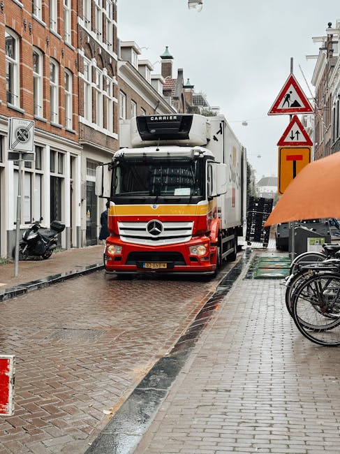 A large Mercedes-Benz moving truck with a white, red, and yellow exterior is positioned on a narrow cobbled street in Holland Park, surrounded by typical Victorian-style terraced residential buildings