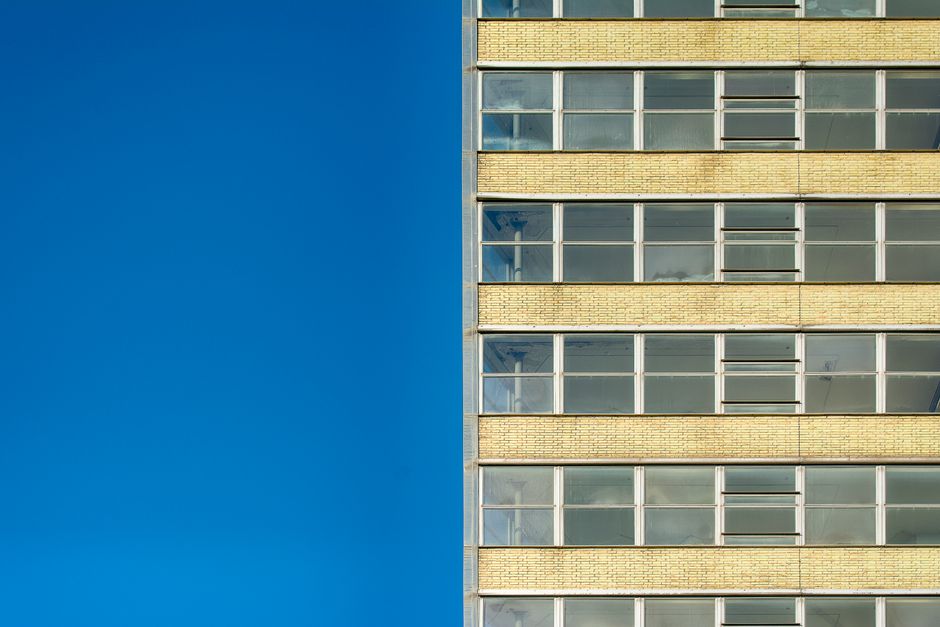 A tall, modern building with multiple stories featuring large rectangular windows arranged in horizontal rows, separated by yellow brickwork. The right side of the image shows the building's yellow brick exterior and glass windows, some of which are open or partially covered, while the left side displays a clear, bright blue sky. This image captures the exterior façade of a multi-storey residential or commercial property, illustrating potential challenges for home relocation or furniture transport services in high-rise access environments, as managed by companies like Man and a Van Holland Park. The scene emphasizes the building's height and window arrangement, relevant to planning efficient packing, loading, and moving logistics for house removals within such structures.