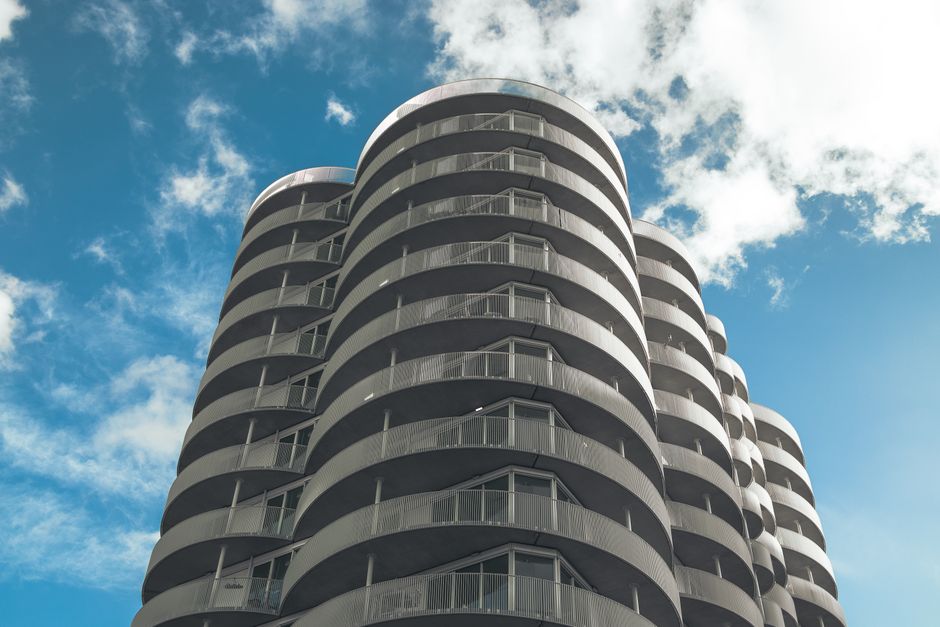 A multi-storey modern residential building with a distinctive curved design, featuring numerous balconies with metal railings, set against a partly cloudy blue sky. The building appears to be a high-rise located in an urban area, with the exterior made of light-colored materials. The image captures the entire height of the structure from a low angle, emphasizing its rounded architectural features. The surrounding environment is not visible, but the building likely presents logistical challenges for home relocations and furniture transport, especially when accessing upper floors. As demonstrated by the exterior design, effective packing and careful loading are essential during moving processes, which is service often managed by companies such as Man and a Van Holland Park, known for handling complex removals including high-rise access problems.