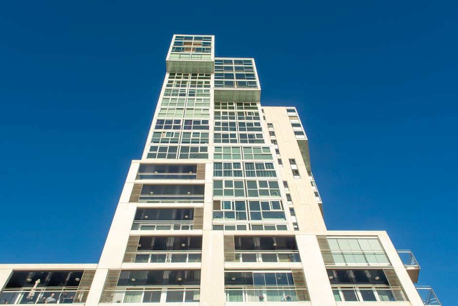 A tall, modern building with multiple stories featuring large rectangular windows arranged in horizontal rows, separated by yellow brickwork. The right side of the image shows the building's yellow brick exterior and glass windows, some of which are open or partially covered, while the left side displays a clear, bright blue sky. This image captures the exterior façade of a multi-storey residential or commercial property, illustrating potential challenges for home relocation or furniture transport services in high-rise access environments, as managed by companies like Man and a Van Holland Park. The scene emphasizes the building's height and window arrangement, relevant to planning efficient packing, loading, and moving logistics for house removals within such structures.