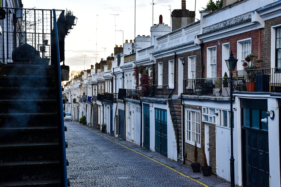 A row of terraced houses in the Leighton House area of Holland Park, with front gardens, potted plants, and decorative lampposts along the pavement. The houses feature brick and painted exteriors, with some having external staircases and metal railings leading to upper-level entrances. The street is cobbled, and the scene is illuminated by natural daylight, suggesting a typical urban setting suitable for home relocation or furniture transport services. This environment exemplifies the type of residential property commonly serviced by Man and a Van Holland Park during packing and moving projects, emphasizing the logistical context of house removals in a historic and dense urban neighbourhood.
