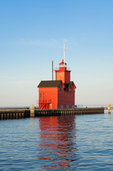 A red wooden lighthouse with small windows and a black roof is situated on a stone pier extending into a calm body of water. The lighthouse has a vertical antenna on top and a staircase leading up to a doorway near the waterline. In the background, the sky is clear with a subtle gradient from light blue near the horizon to a slightly darker blue higher up, indicating daytime. The water reflects the lighthouse and sky, creating a mirror-like effect. This scene is outside a building or property for house removals, showcasing the type of environment where furniture transport and home relocation services by Man and a Van Holland Park might operate, especially when moving items across water or along coastal routes.