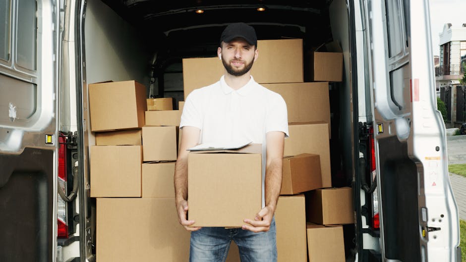 A man wearing a white polo shirt, blue jeans, and a black cap stands inside an open van during a home relocation process, holding a medium-sized cardboard box with both hands. The van's interior is filled with stacked cardboard boxes of various sizes, some wrapped in plastic, ready for loading or unloading. The van is parked outdoors on a pavement near residential buildings, with sunlight illuminating the scene. The image captures the loading or packing stage of furniture transport by Man and a Van Holland Park, highlighting careful handling of boxes during the moving process, including the use of cardboard packaging and the vehicle prepared for house removals and urgent moves.