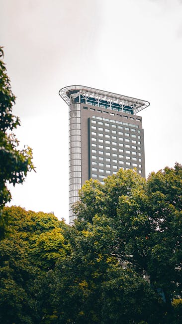 A tall, modern skyscraper with a rectangular shape and numerous evenly spaced windows, standing prominently above a cluster of green trees with dense foliage in the foreground. The building features a distinctive curved, overhanging roof at the top, supported by exposed structural elements, and appears to be an office or commercial tower. The sky above is overcast with gray clouds. This image is relevant to house removals and furniture transport services offered by Man and a Van Holland Park, illustrating a typical urban setting where home relocation and moving logistics may take place, with the building's height and structure emphasizing the scale of property and furniture moving operations in busy city environments.